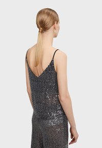 Tmavošedý top bez rukávov vyrobený z flitrového materiálu, s dizajnom V-zadnej časti a tenkými ramienkami. Flitre odrážajú svetlo.