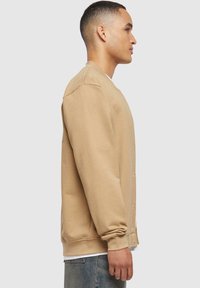 Jeune homme en sweat beige boutonné et jean bleu, debout de profil sur un fond clair uni.