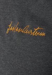 Grauer Stoff mit einer feinen Textur zeigt goldgelbe gestickte Schriftzüge in Kursiv, die prominent auf der Oberfläche platziert sind.