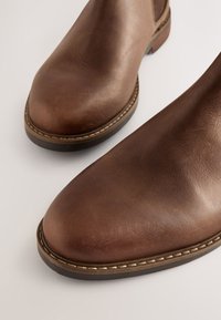 Bottes chevilles en cuir marron avec un bout arrondi, une semelle plate et des détails de couture minimaux le long du bord. Texture lisse et design classique.