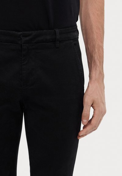 Pantalons en coton noir à couper droit, avec une texture douce, des passants de ceinture et des poches latérales avant. Main partiellement visible sur le côté.