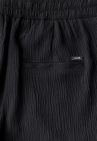 Tejido negro texturizado con un diseño arrugado, con una banda elástica en la cintura y una pequeña etiqueta metálica de la marca en el bolsillo trasero.