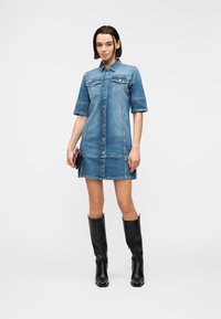 Νεαρή γυναίκα με κοντομάνικο, κουμπωτό φόρεμα από τζιν, κρατώντας μια τσάντα clutch και φορώντας μαύρες μπότες μέχρι τη μέση της γάμπας, σε λευκό φόντο.