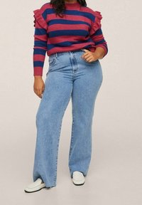 Pullover mit roten und blauen Streifen, ausgestattet mit Rüschendetails an den Ärmeln. Hochgeschnittene blaue Jeans mit weitem Bein. Weiße Slip-On-Schuhe.