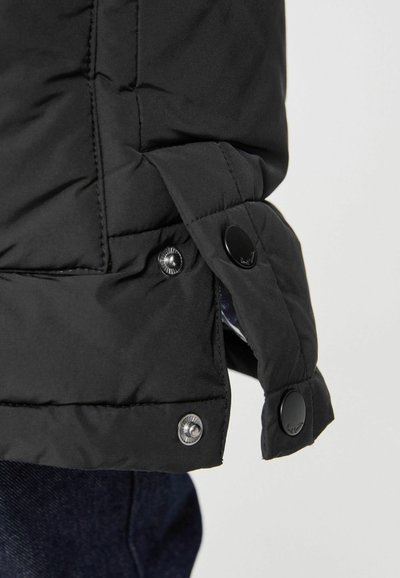 Melnā izolētā jaka ar quilted tekstūru, ar pogām un nedaudz paplašinātu apakšdaļu, kas paredzēta siltumam un funkcionalitātei.