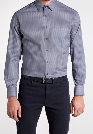 Homme portant une chemise ajustée à manches longues à motifs, boutonnée et rentrée dans un pantalon foncé avec une ceinture noire, debout devant un fond blanc.