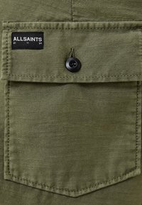 Tessuto verde oliva sovrapposto con tasca a pattina e bottone nero. Include un'etichetta nera con testo bianco "ALLSAINTS" e dettagli numerici.