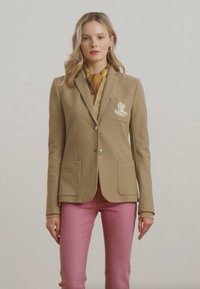Beige lyhyt blazer, jossa on brodeerattu logo taskussa, kaksi etutaskua ja napitus, yhdistettynä vaaleanpunaisiin housuihin ja vaaleaan paitaan.