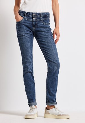 Jeans in denim con lavaggio blu medio, slim fit, design a cinque tasche e chiusura a doppio bottone. Orli arrotolati e abbinati a sneakers bianche.