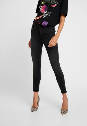 Czarne obcisłe dżinsy noszone z czarnym koszulkiem z grafiką kolorowej rakiety i planet, połączone z wysokimi obcasami w wzór skóry węża.