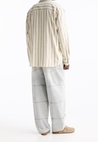 Chemise à rayures beiges avec une coupe décontractée, associée à un pantalon gris clair à jambes larges avec des coutures visibles et des poches. Texture douce.