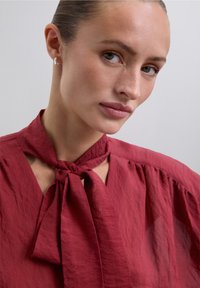 Blusa roja con textura y cuello atado. Presenta un corte ligeramente holgado y un escote abierto para un estilo relajado. Pendientes de aro de plata simples.