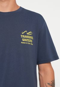 Marineblå bomulds t-shirt med et gult grafisk design, der står "TRANQUIL WATERS" og "Relax to the Max" på brystet.