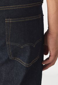 Mörka denimjeans med en ficka som har kontrasterande gul söm och en distinkt böjd design. Närbild på fickan och handen.