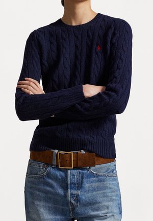 Osoba ubrana w granatowy sweter z grubym splotem z czerwonym logo, niebieskie dżinsy i brązowy skórzany pasek, z założonymi na piersi skrzyżowanymi ramionami.