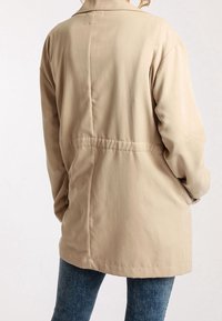 Chaqueta beige de longitud media con un ajuste relajado, que presenta un cuello con solapa, costuras visibles y una tela que cae suavemente.