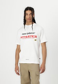 Balts kokvilnas T-krekls ar melno un sarkano "new balance ATHLETICS" apdruku, ar mazu logotipa detaļu uz krūtīm un tekstu apakšā.