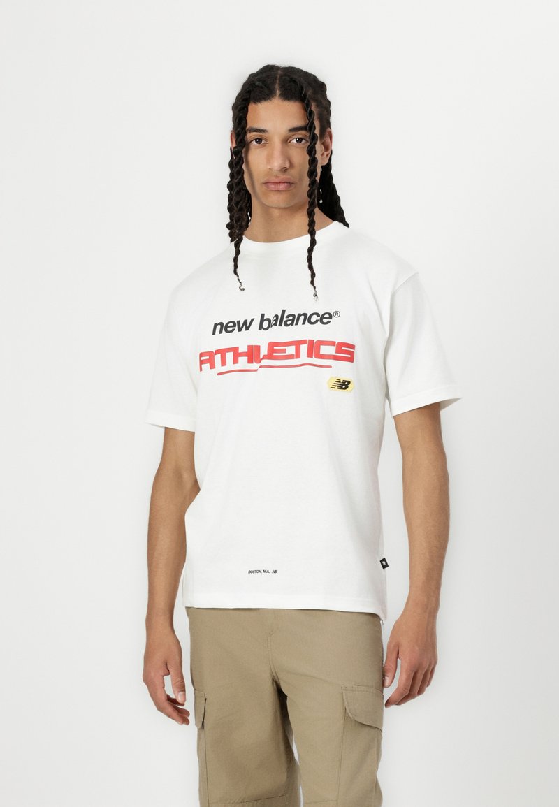 Balts kokvilnas T-krekls ar melno un sarkano "new balance ATHLETICS" apdruku, ar mazu logotipa detaļu uz krūtīm un tekstu apakšā.