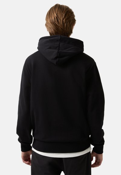 Persoon met lichtbruin haar die een zwarte hoodie over een wit shirt draagt, staande met hun rug naar de kijker gekeerd.