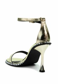 Złote sandały na wysokim obcasie z paskiem wokół kostki, smukłym obcasem typu stiletto i ozdobami z kryształków wzdłuż krawędzi podeszwy.
