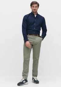 Marinblå skjorta med knappar, ljusgröna byxor, vävd brun skärp och marinblå sneakers med vita accenter. Modell som står i en neutral bakgrund.