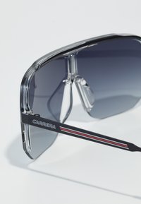 Occhiali da sole con montatura trasparente e lenti grigio sfumate. Le astine nere presentano strisce rosse e bianche, con il logo "CARRERA".