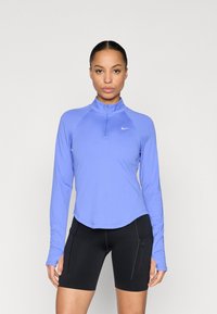 Top deportivo morado de manga larga con media cremallera y dobladillo redondo, hecho de tela elástica, combinado con pantalones cortos ajustados negros con bolsillos con cremallera.