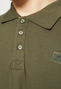 Olivově zelená polo košile s límečkem, třemi knoflíky a malou nášivkou s logem. Tkanina je hladká s viditelnými švy.