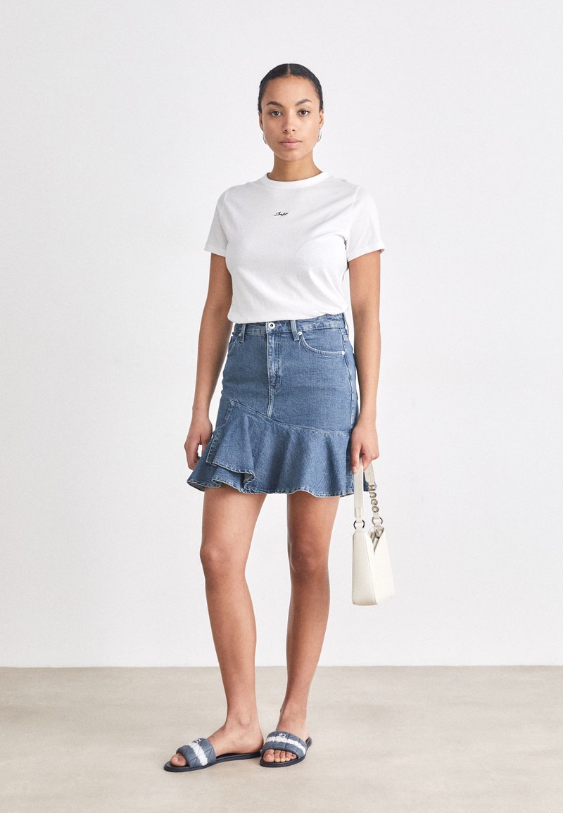T-shirt blanc avec texte brodé, jupe en denim bleu avec un ourlet volanté, sandales à motif et petit sac à main blanc. Fond uni.