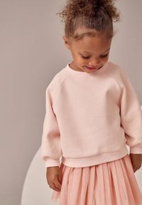 Sweat-shirt rose clair avec manches raglan et poignets côtelés, associé à une jupe rose transparente à volants. Textures douces et design simple.