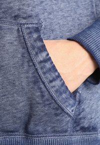 Blå huvtröja med borstad tygtextur, ribbade ärmar och en känguruficka på framsidan, med en lätt blekt effekt.