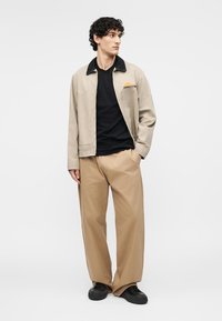 Jeune homme aux cheveux bouclés portant une veste beige avec un col noir, une chemise noire, un pantalon beige à jambes larges et des chaussures noires, debout avec une main dans la poche.