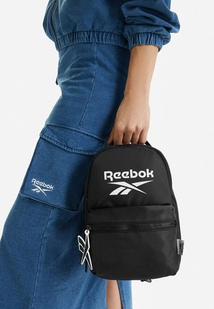 Persona con conjunto de mezclilla azul sosteniendo una pequeña mochila negra de Reebok por su asa superior contra un fondo blanco liso.