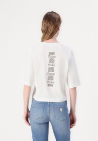 T-shirt bianca corta con maniche corte, che presenta un design verticale ripetitivo del logo "Pinko" in grigio sul retro. Indossata con jeans azzurri chiari.