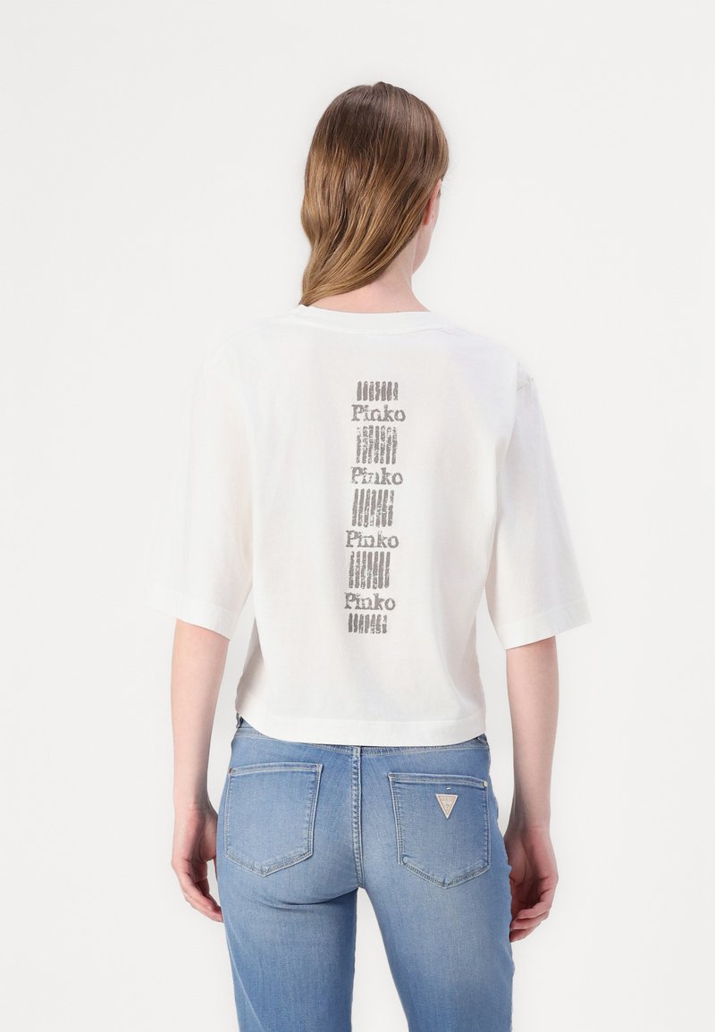 T-shirt bianca corta con maniche corte, che presenta un design verticale ripetitivo del logo "Pinko" in grigio sul retro. Indossata con jeans azzurri chiari.
