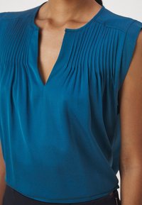 Blouse sans manches bleu sarcelle avec un col en V et des plis verticaux sur la poitrine, portée par une personne à la peau brune.