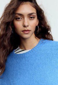 Hellblauer Strickpullover mit geripptem Rundhalsausschnitt, darüber ein schwarz-weiß gestreiftes Oberteil getragen. Glatte Textur, lockere Passform.