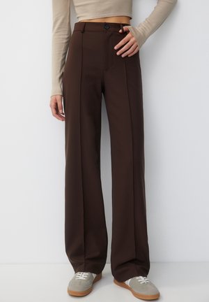 Femme portant un pantalon marron foncé taille haute à jambes larges avec un bouton à l'avant et un crop top beige à manches longues, debout contre un mur blanc.