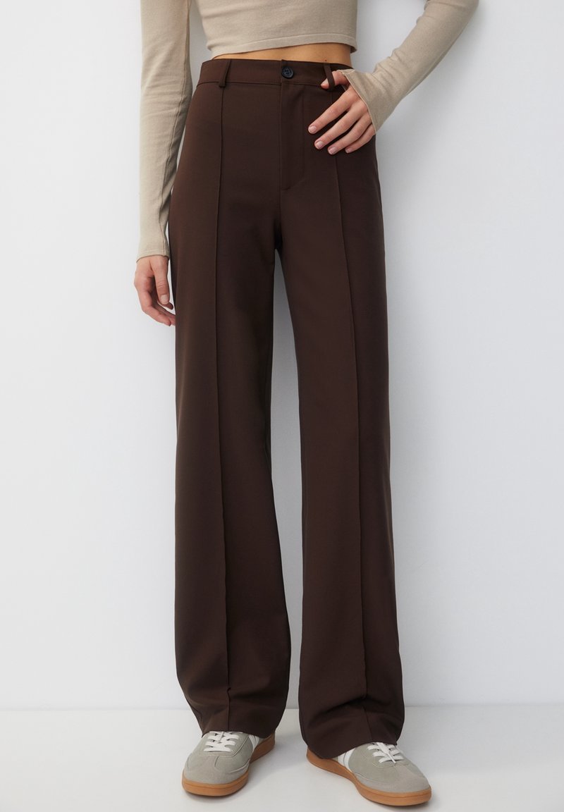 Femme portant un pantalon marron foncé taille haute à jambes larges avec un bouton à l'avant et un crop top beige à manches longues, debout contre un mur blanc.