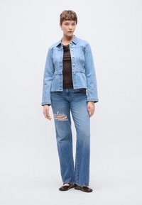 Lichtblauwe spijkerbroekjas met een getailleerde taille, knoopsluiting en lange mouwen, gecombineerd met versleten blauwe spijkerbroeken en bruine platte schoenen.