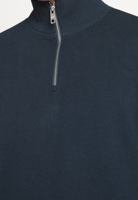 Marineblauw ritspullover met hoge kraag, geribbelde textuur en zilveren rits. Close-up weergave concentreert zich op de halslijn en stiksels.