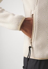 Main à la peau foncée dans la poche latérale zippée d'une veste texturée beige à motif avec des cordons de fermeture éclair noirs.