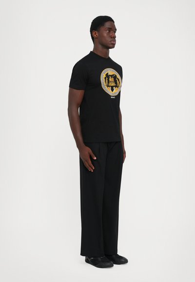 Bărbat îmbrăcat cu un tricou negru Versace cu emblema aurie, pantaloni largi negri și pantofi negri, stând pe un fundal alb.