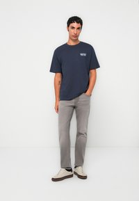 Joven vestido con una camiseta gráfica azul marino, jeans gris claro y zapatillas beige, de pie contra un fondo blanco liso.