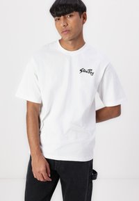 White cotton t-shirt with short sleeves, featuring a black logo printed on the left chest. Paired with dark jeans.