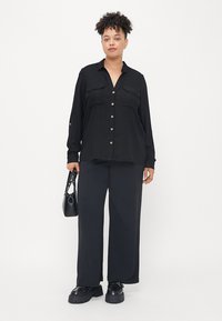 Chemise noire à manches longues avec boutons et deux poches poitrine, assortie à un pantalon noir à jambes larges et des chaussures noires épaisses. Les accessoires incluent un petit sac noir.