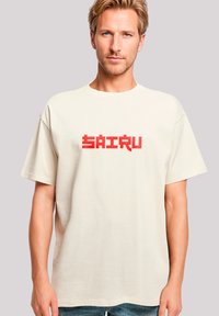 Beige cotton t-shirt with short sleeves, featuring a bold red pixelated "SAIRU" text graphic centered on the front. Casual fit.