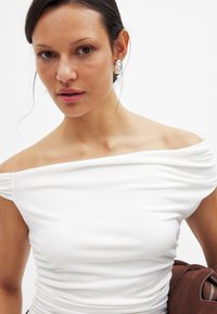 White off-the-shoulder top with ruching detail, smooth texture, and fitted design. Model wears large decorative earrings and holds a brown garment.