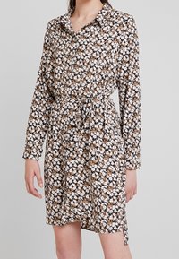 Blommig skjortklänning med långa ärmar, krage och dragsko i midjan. Tyget har en svart bakgrund med vita och bruna blommor.