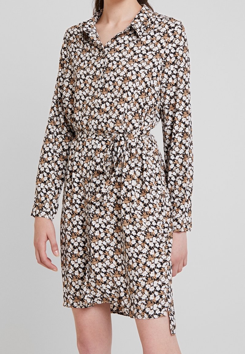 Blommig skjortklänning med långa ärmar, krage och dragsko i midjan. Tyget har en svart bakgrund med vita och bruna blommor.